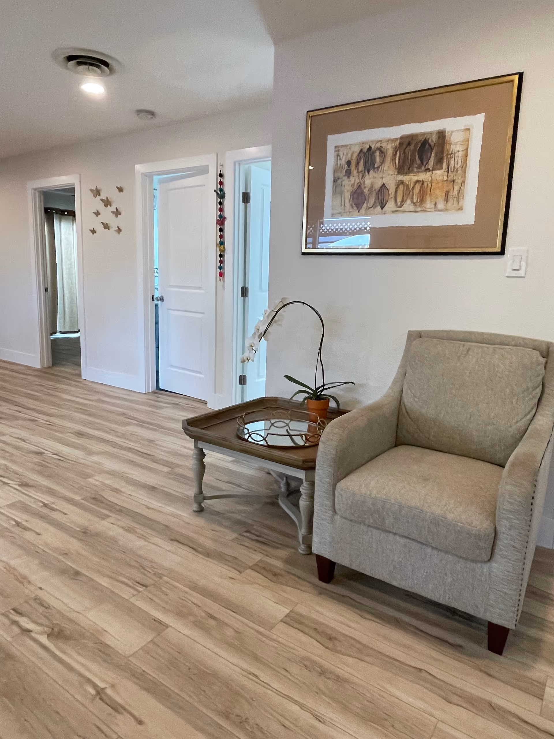 A cozy interior space with light wood flooring, a beige upholstered armchair, and a wooden side table holding a potted orchid plant. On the wall above the chair is a framed abstract artwork. In the background, there are two white doors and a hallway with butterfly wall decorations.