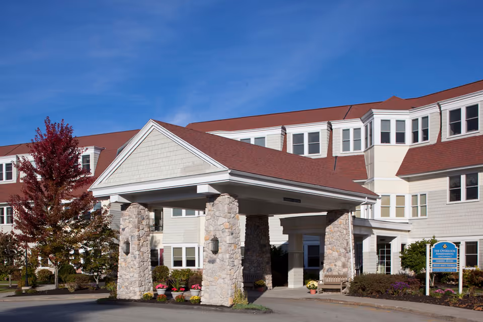 Front entrance of a senior living facility featuring a covered drive-through with stone columns and a red-roofed multi-story building.