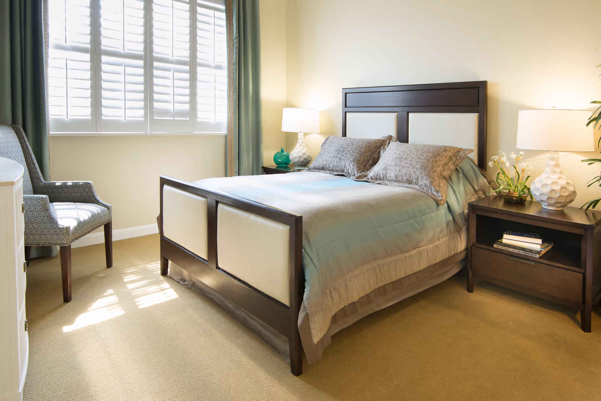 Sunlit bedroom with a made bed featuring a dark wood headboard, two nightstands with lamps, an upholstered chair, and a window with shutters.
