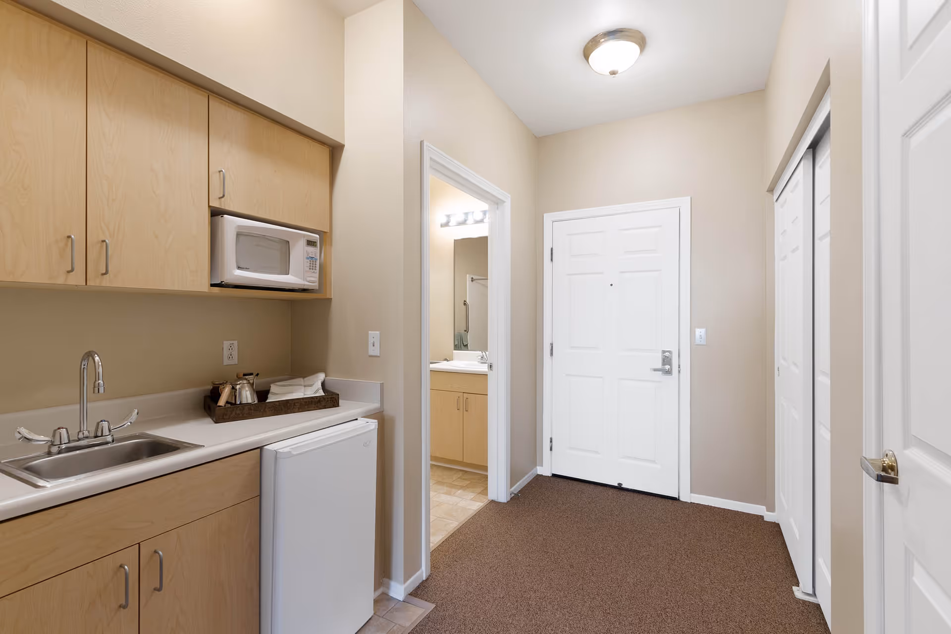 Interior view of a small kitchenette area with light wood cabinets, a microwave, a sink, and a mini refrigerator. Adjacent to the kitchenette is a hallway with a white door and a bathroom visible through an open doorway. The walls are painted beige and the floor is carpeted in the hallway area.