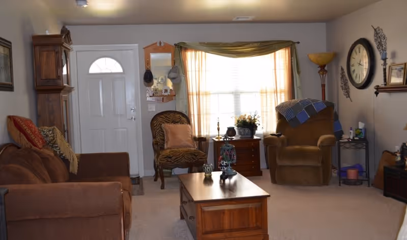 A cozy living room with a brown sofa on the left, a patterned armchair and a brown recliner chair near a window with sheer curtains. A wooden coffee table is centered on a beige carpet. The walls are decorated with a large clock, a floor lamp, and small shelves with decorative items. The white front door is visible in the background.