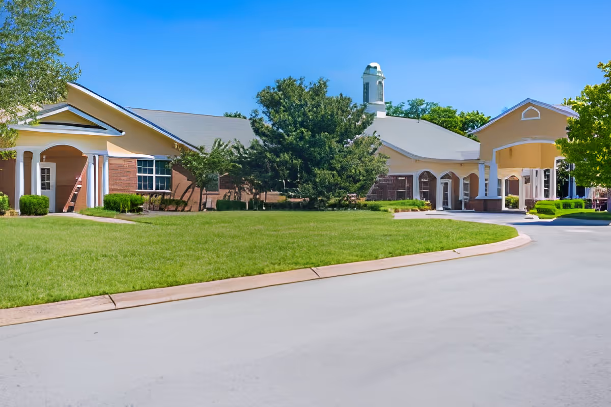 Exterior view of a senior living facility building with a driveway, green lawn, trees, and clear blue sky.