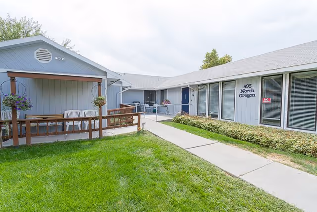Exterior view of Ashley Manor Senior Living showing a single-story building with gray siding, multiple windows with blinds, a blue door, and a covered porch area with white chairs. A concrete walkway leads through a green lawn with some shrubs along the building.