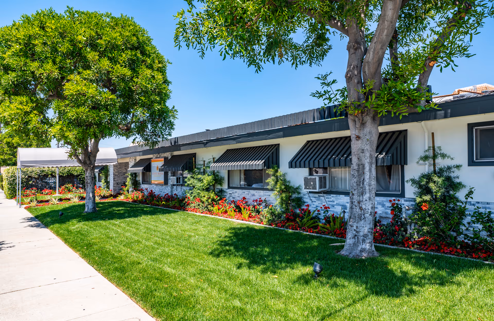 Exterior view of a single-story building with black awnings over the windows, surrounded by green grass, trees, and flower beds under a clear blue sky.