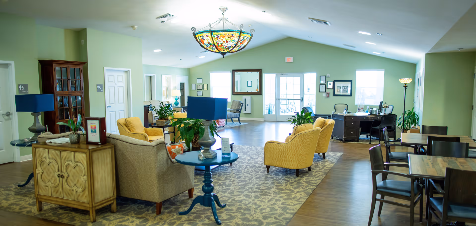 A spacious and well-lit common area in a senior living facility with green walls, a patterned carpet, and wooden flooring. The room features several yellow and beige armchairs arranged around blue side tables with lamps and plants. There are multiple framed pictures and a large mirror on the far wall, along with a desk and chairs near the windows and door. A colorful stained glass chandelier hangs from the ceiling.