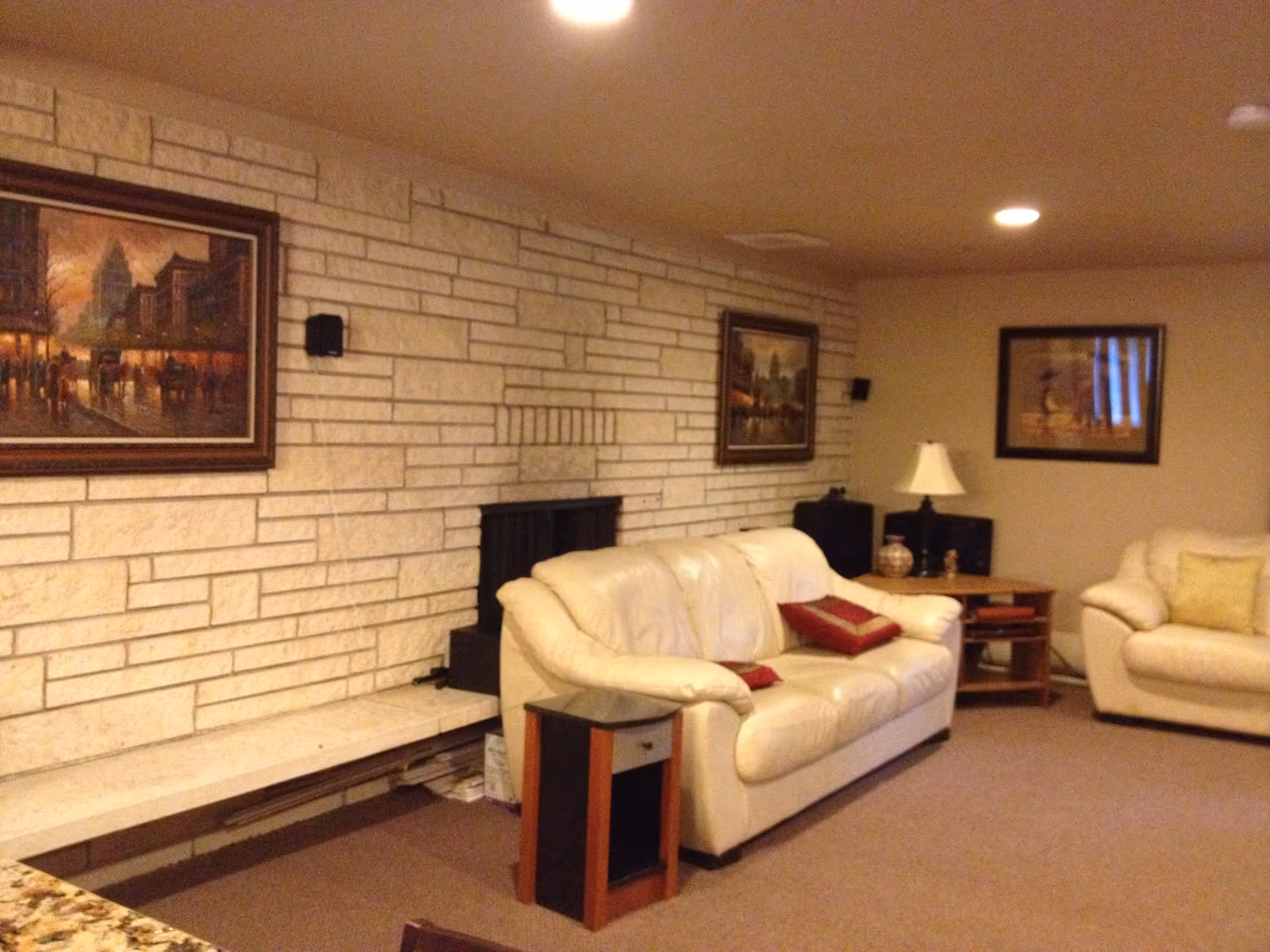 A cozy living room with beige leather sofas, a small wooden side table with a lamp, and framed paintings on a light-colored brick wall. The room has a carpeted floor and recessed ceiling lights.