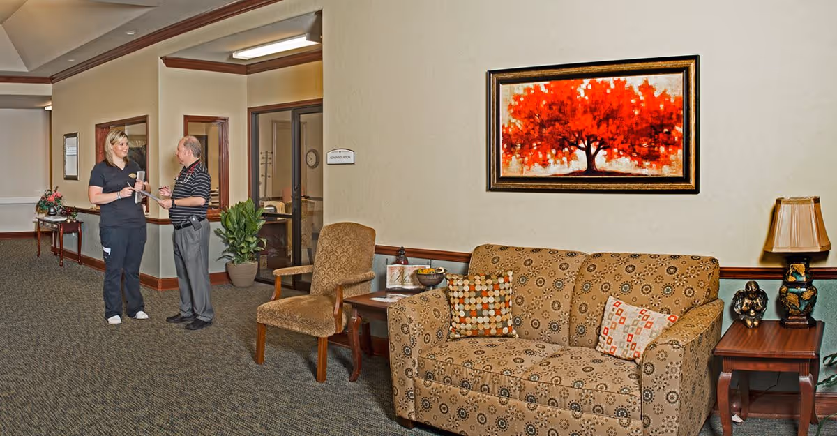 A hallway in a senior living facility with a patterned sofa and armchair, a side table with a lamp and decorative items, and a large framed painting of a red tree on the wall. Two people, a woman in a black uniform and a man in a striped shirt, are standing and talking near a doorway labeled 'Administration'.