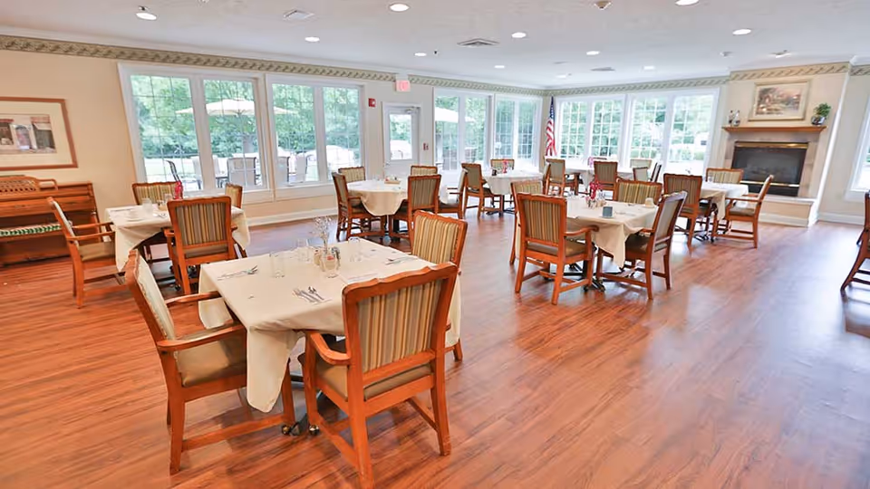 A bright and spacious dining room with multiple tables covered in white tablecloths, each surrounded by wooden chairs with striped cushions. Large windows let in natural light and offer a view of greenery outside. There is a fireplace on the right side of the room and an American flag near the back door.