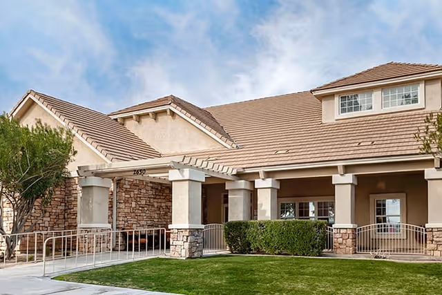 Exterior view of a single-story senior living facility building with beige walls, stone accents, a tiled roof, and a covered entrance supported by columns. There is a well-maintained lawn and a tree in front of the building under a partly cloudy sky.