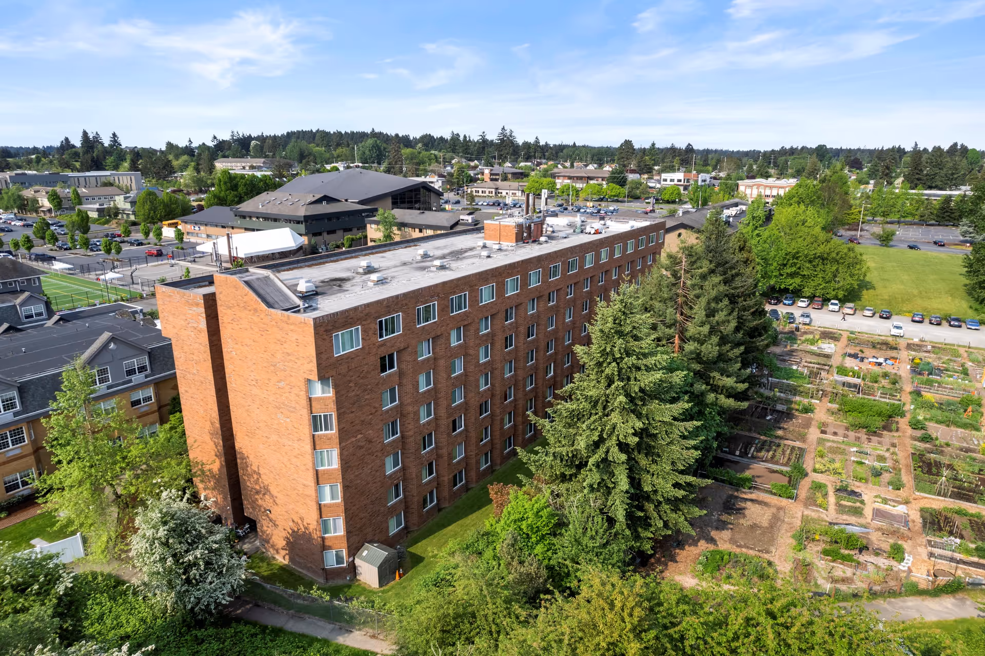 Aerial view of a multi-story brick building surrounded by trees and greenery, with a community garden and parking lot nearby under a partly cloudy sky.
