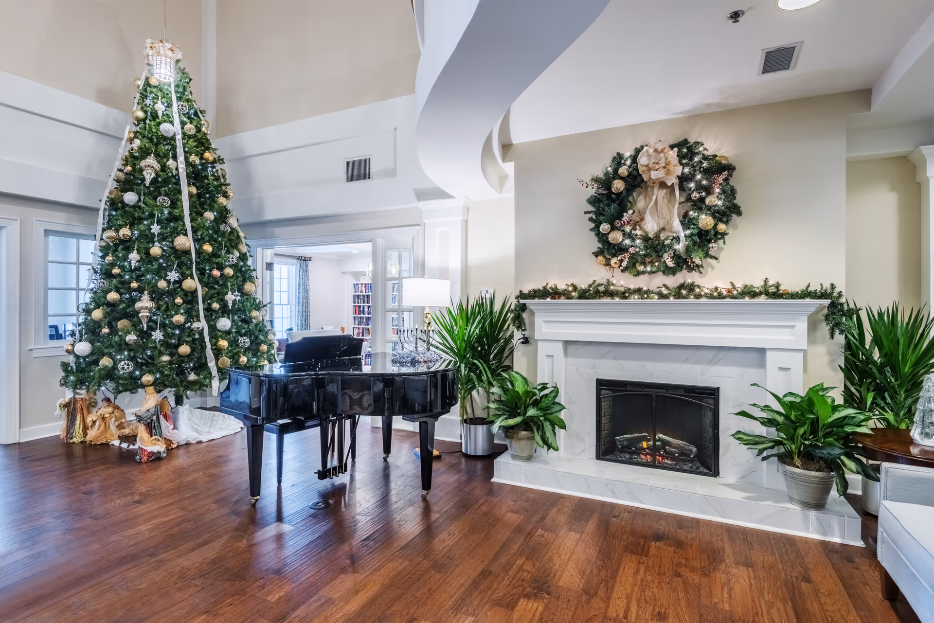 A spacious and elegantly decorated living room area featuring a large Christmas tree adorned with gold and white ornaments, a black grand piano, a white fireplace with a festive wreath above it, and several green potted plants. The room has wooden flooring and light-colored walls with a curved ceiling design.
