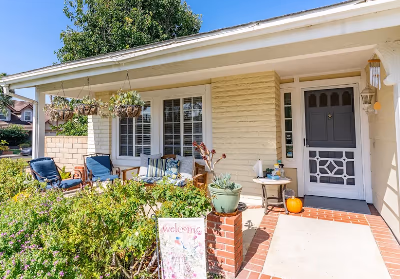Front porch of Irvine Cottage #1 featuring outdoor seating, potted plants, and a welcome sign.