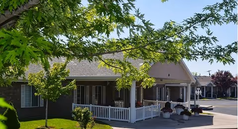 Front exterior of a single-story senior living building with a covered porch, white railing, potted plants and trees in the foreground.
