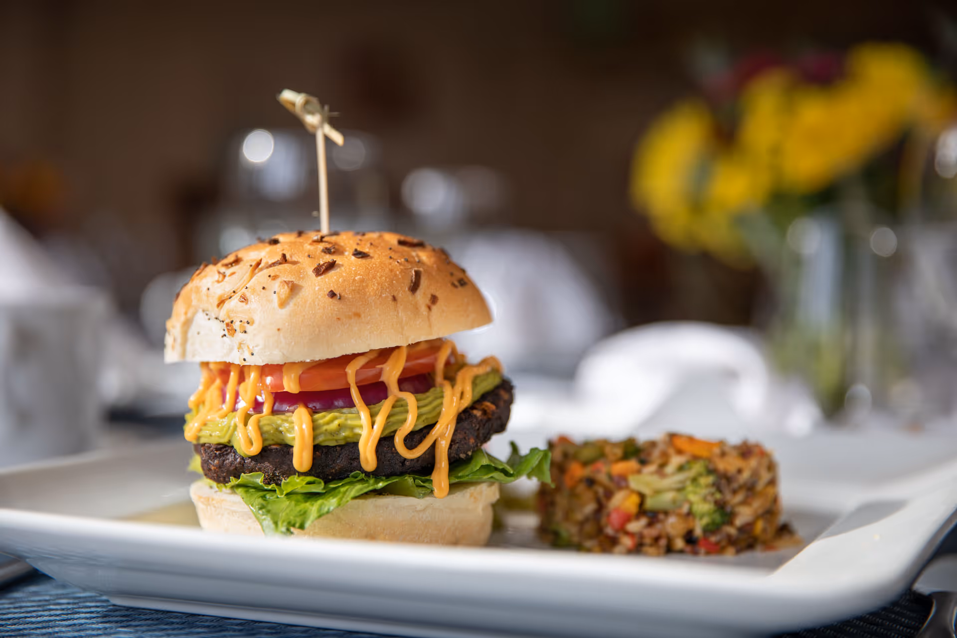 A close-up of a gourmet hamburger with lettuce, tomato, onion, guacamole, and a drizzle of orange sauce on a toasted bun, served on a white plate with a side of mixed vegetables and grains. The background is softly blurred with hints of yellow flowers and table settings.