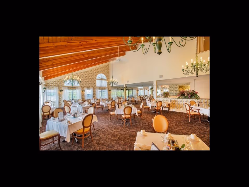 A spacious dining room with multiple tables covered in white tablecloths, each set with cups, glasses, and condiments. The room features high wooden ceilings with exposed beams, large windows with sheer curtains, floral wallpaper, and elegant chandeliers hanging from the ceiling. The chairs have cushioned seats and backs in a warm tan color.