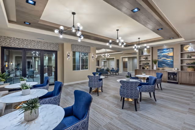 A spacious and well-lit common area in a senior living facility featuring multiple round marble tables with blue upholstered chairs. The room has modern pendant lighting, large windows, and built-in shelves with decorative items and artwork on the walls.