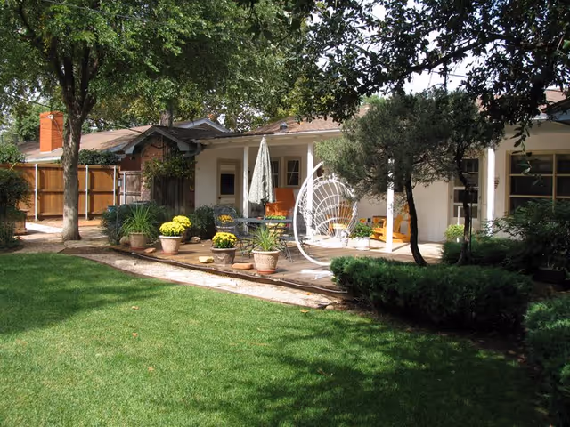 A backyard patio area of a single-story home with a green lawn in the foreground. The patio features potted plants with yellow flowers, a white hanging chair, a table with an umbrella, and various shrubs and trees providing shade.