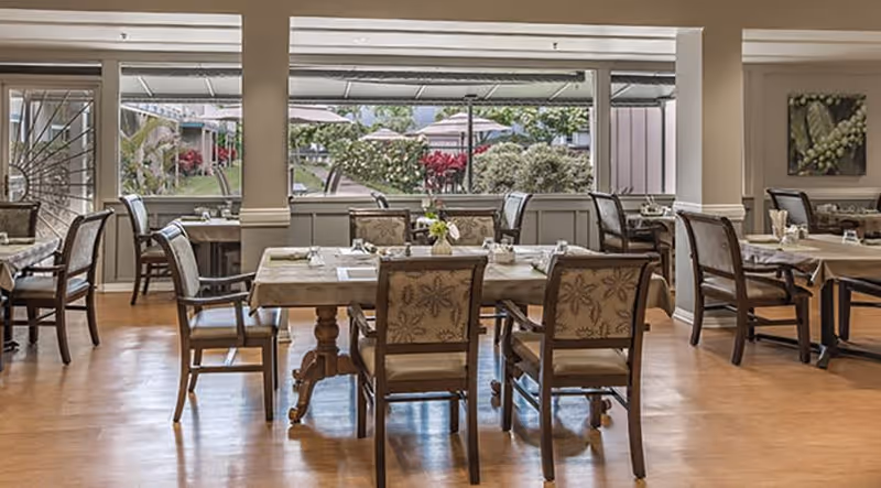 A dining room with several wooden tables and chairs arranged neatly. Each table is set with placemats, napkins, and small flower vases. Large windows in the background provide a view of an outdoor garden area with greenery and red flowers.
