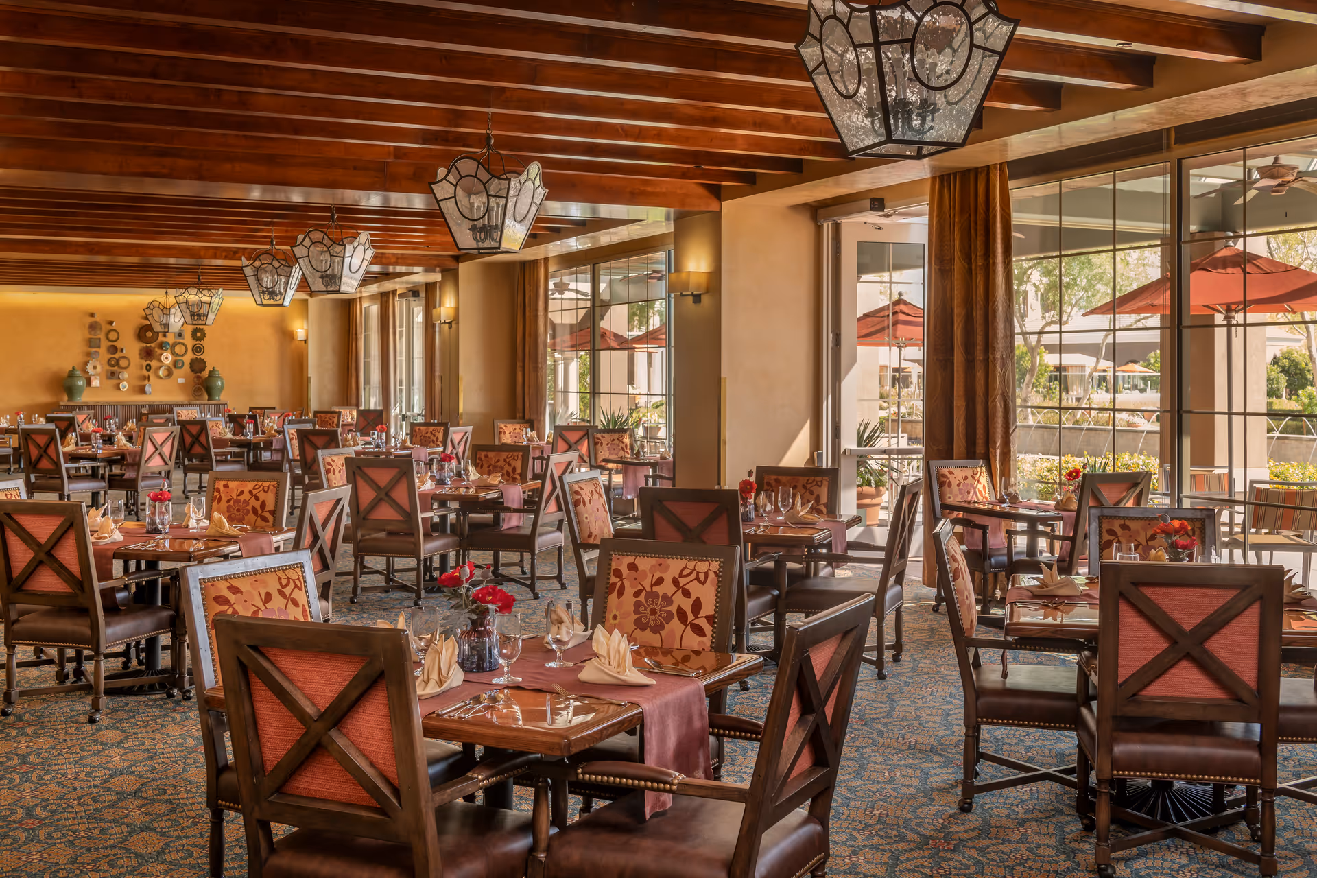 A spacious dining room with wooden beams on the ceiling and large windows letting in natural light. The room is furnished with multiple wooden tables and chairs, each table set with folded napkins, glassware, and small vases with red flowers. Outside the windows, red umbrellas and greenery are visible.