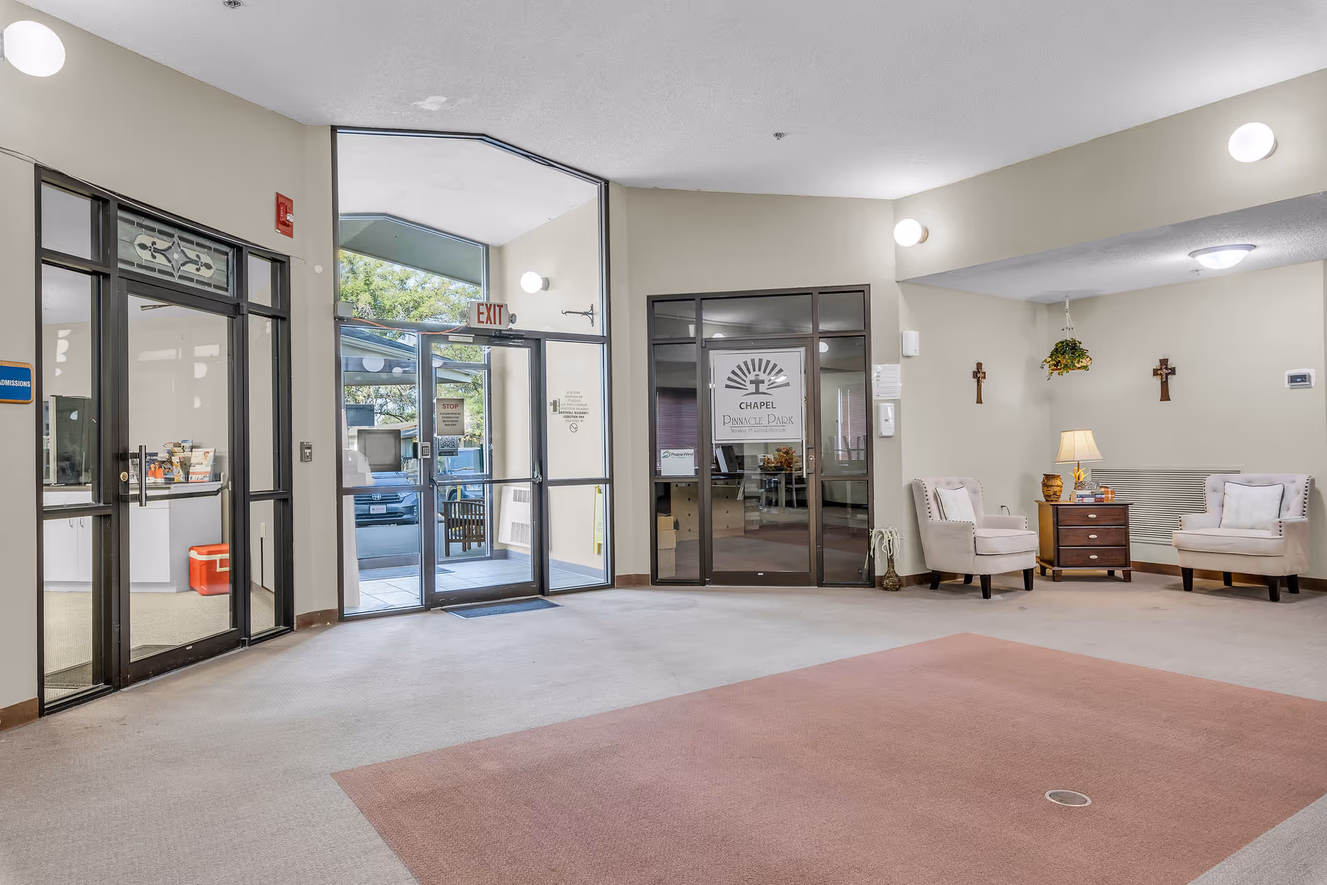 Interior view of a senior living facility lobby area with beige walls and carpet. There are two beige armchairs with white pillows, a wooden side table with a lamp and decorative items, and two crosses hanging on the wall. Glass doors lead to an outside exit and to a chapel room labeled 'Chapel Pinnacle Park'.