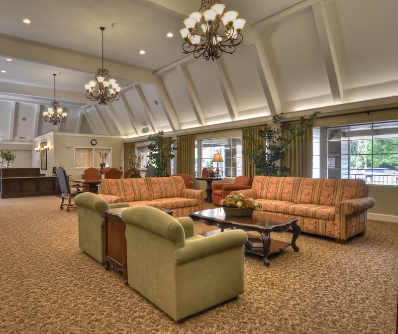 A spacious and well-lit living room area with high vaulted ceilings and multiple chandeliers. The room features patterned sofas, green armchairs, a wooden coffee table with a decorative centerpiece, and large windows with curtains allowing natural light. There are also plants and additional seating areas visible in the background.