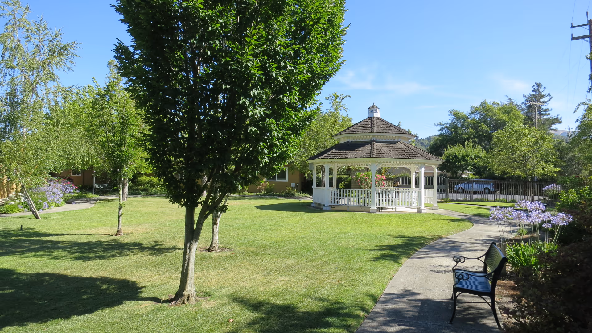 A sunny outdoor garden area at Creekwood Care Facility featuring a white gazebo with a shingled roof, surrounded by green grass, trees, and flowering plants. A curved concrete pathway leads to the gazebo, with a black metal bench positioned along the path. The sky is clear and blue.