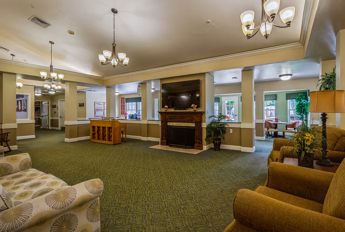 Spacious senior living common area with upholstered seating, a fireplace and TV, a small piano, and dining tables visible in the background.