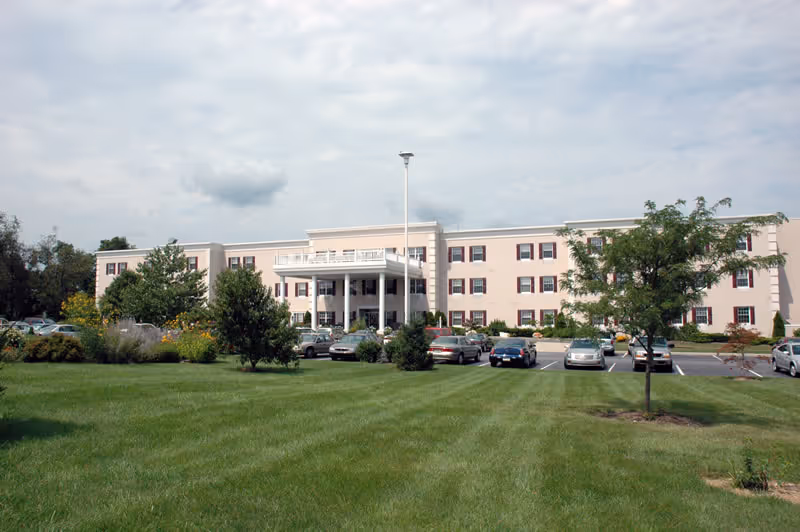 Front exterior view of a three-story beige retirement community building with multiple windows and a covered entrance. There is a parking lot with several cars in front, surrounded by green grass, trees, and shrubs under a cloudy sky.