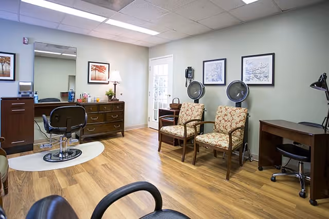 Interior view of a salon area in a senior living facility with wooden flooring, two salon chairs, a dresser with a lamp and mirror, two hair drying stations with patterned chairs, framed artwork on the walls, and a desk with a lamp and chair.
