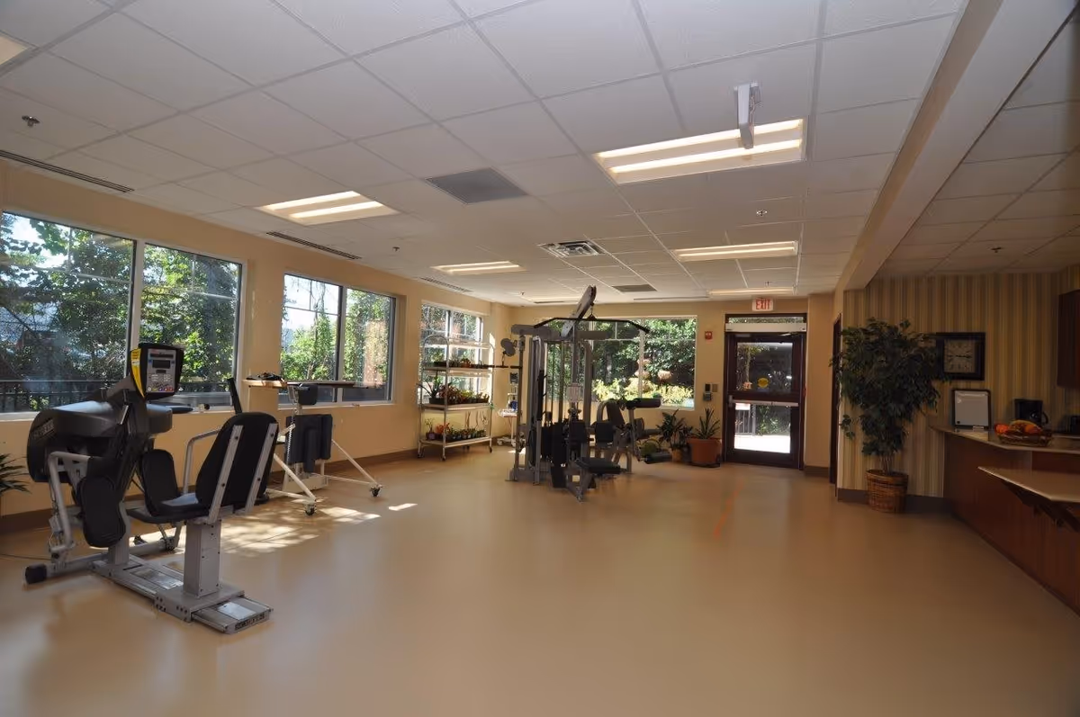 A spacious fitness room with exercise equipment including a leg press machine and a multi-station gym. Large windows on the left side let in natural light and show greenery outside. There is a door at the far end leading outdoors, and a small counter area with a plant and a clock on the right side.