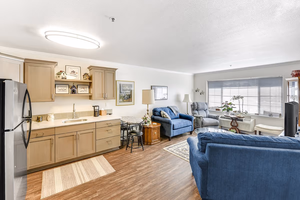 A bright and spacious living area featuring a small kitchen with light wood cabinets, a stainless steel refrigerator, and a sink. Adjacent to the kitchen is a small dining table with two chairs. The living room area includes two blue upholstered sofas, a gray recliner chair, a wooden side table with a lamp, and a large window with blinds allowing natural light to fill the room. The floor is wood with a small rug near the kitchen area.