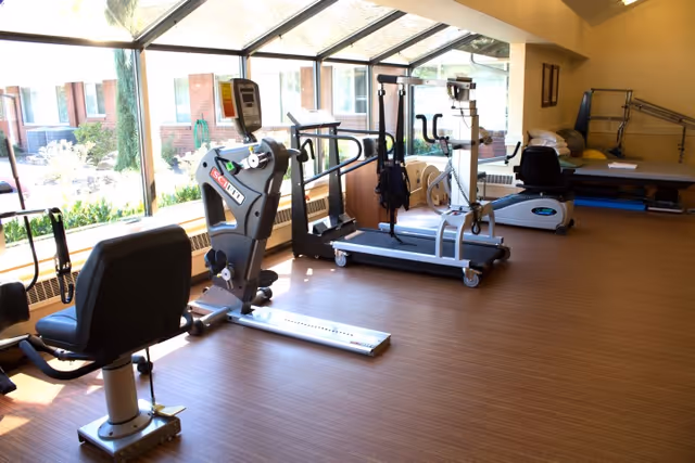 A bright exercise room with large windows showing an outdoor view. The room contains various fitness equipment including stationary bikes, a treadmill, and other exercise machines. The floor is wooden, and there are exercise mats and balls in the background.