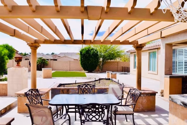 Outdoor patio area with a pergola overhead, a glass-top table surrounded by six metal chairs with cushions, a built-in stone bench, and a basketball hoop attached to the pergola. In the background, there is a green lawn, trees, and a beige fence with neighboring houses visible beyond.