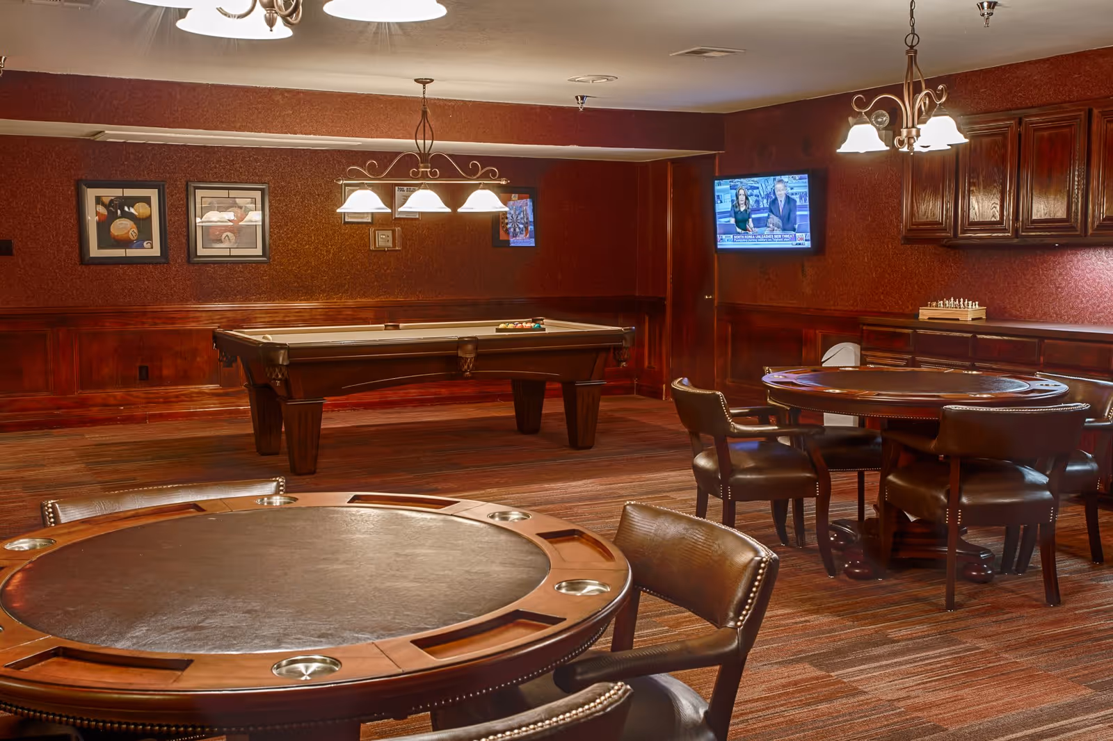 A cozy game room with a pool table in the center, two round card tables with leather chairs, a wall-mounted TV showing a news program, and wooden cabinets along the wall. The room has warm lighting and framed pictures on the walls.