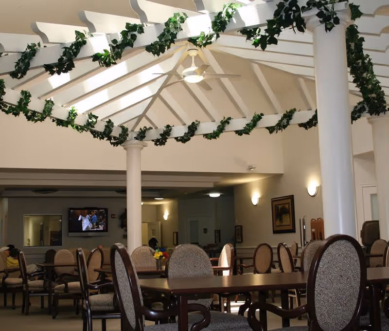 Interior view of a common area with multiple round tables and cushioned chairs arranged for seating. The ceiling features white beams decorated with green leafy garlands and a ceiling fan in the center. The walls have framed artwork and wall-mounted lights, and a television is visible in the background.