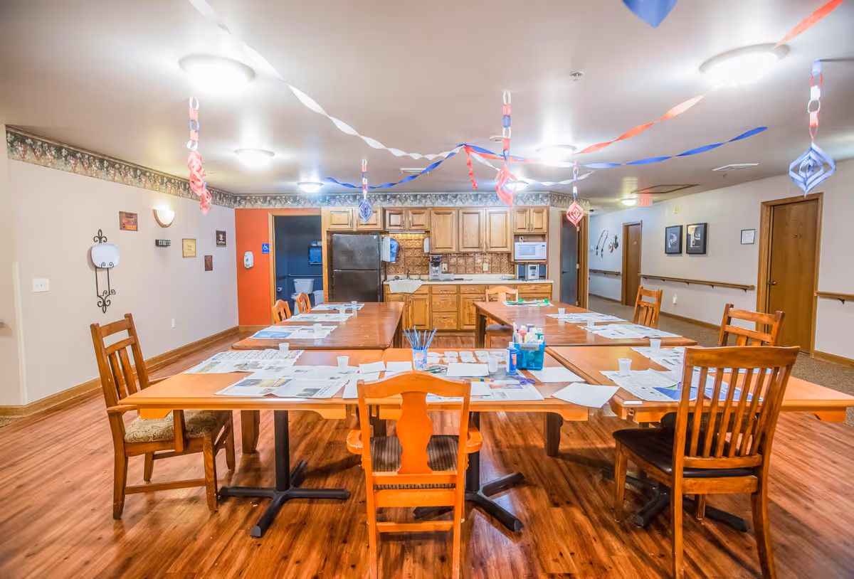 A communal dining/activity room with long tables and wooden chairs arranged for crafts and a kitchenette in the background.