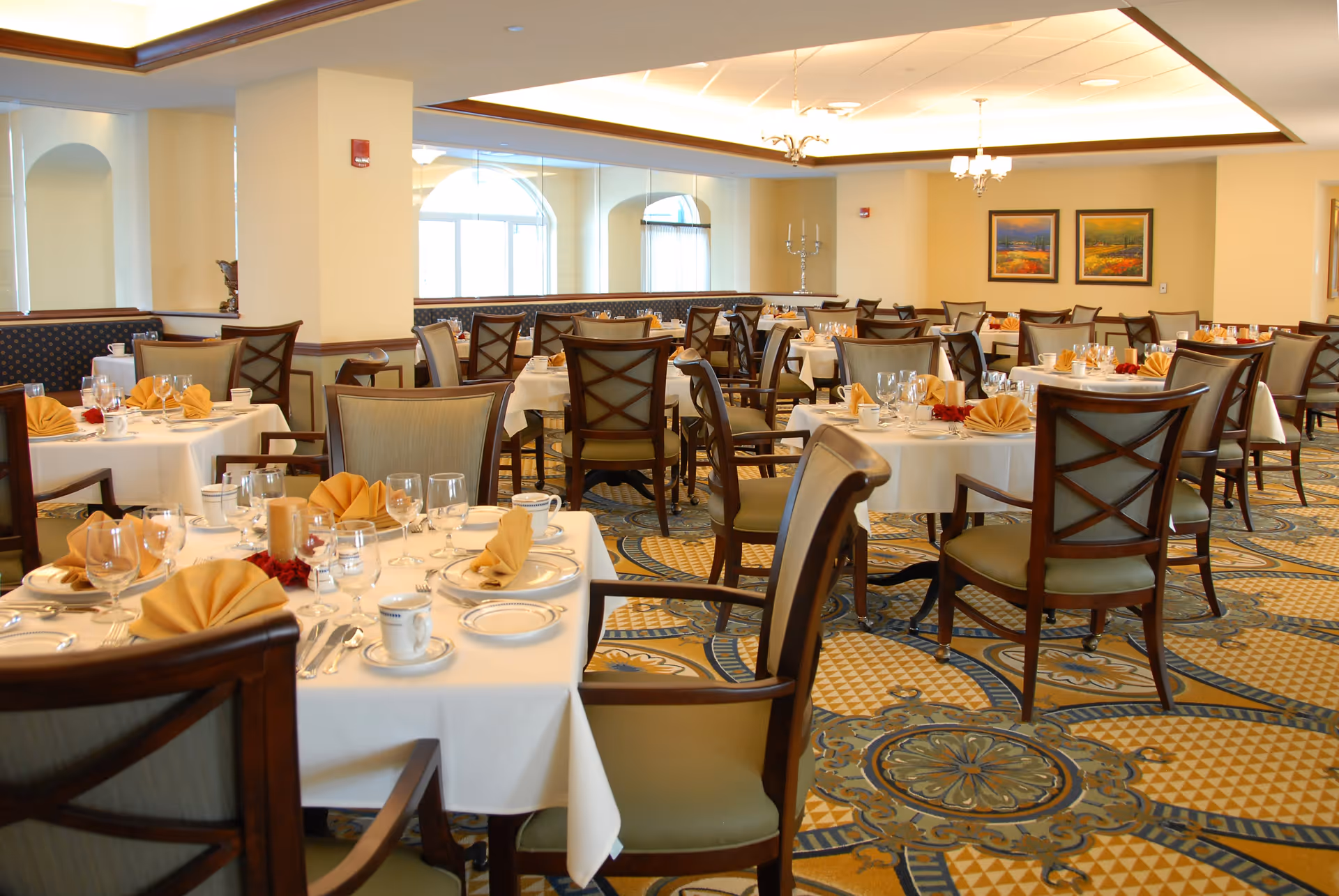 A spacious dining room with multiple tables set with white tablecloths, folded beige napkins, plates, cups, and glassware. The room features wooden chairs with cushioned seats, patterned carpet, and framed artwork on the walls. Large windows allow natural light to brighten the space.