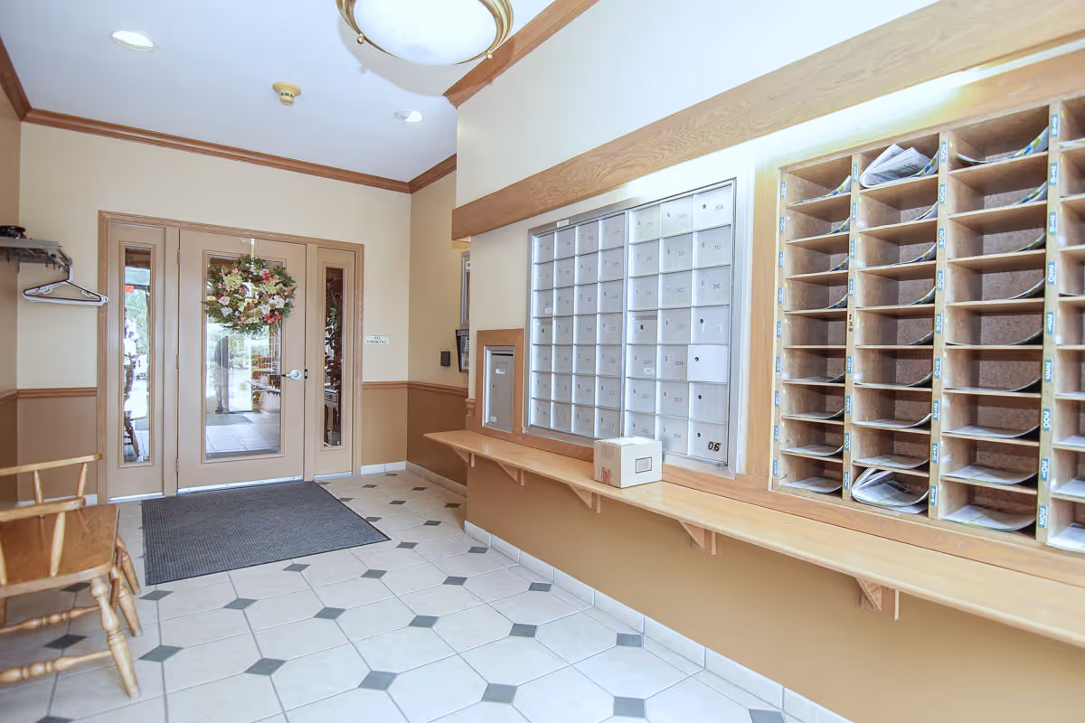 Interior view of a senior apartment mailroom with multiple mailboxes and cubbyholes filled with mail. There is a wooden bench along the wall beneath the mailboxes, a door with glass panels decorated with a wreath, a coat rack with a hanger, and a wooden chair on the left side. The floor is tiled with a geometric pattern.
