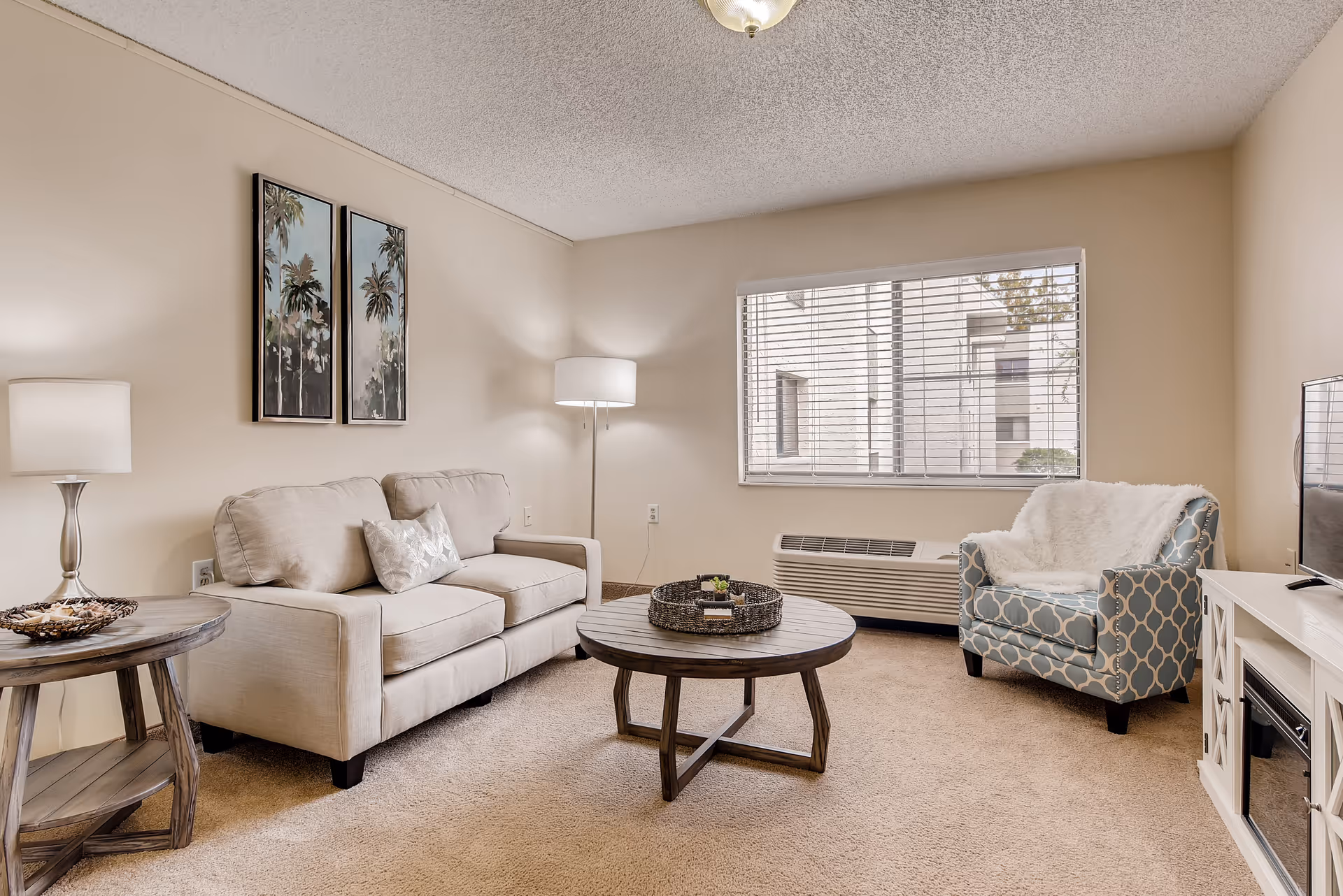 A cozy living room with beige walls and carpet. There is a beige sofa with a decorative pillow, a round wooden coffee table with a woven tray and small plants, a patterned blue armchair with a white throw blanket, a white TV stand with a flat-screen TV, a floor lamp, a table lamp on a round wooden side table, and two framed pictures of palm trees on the wall. A large window with blinds lets in natural light.