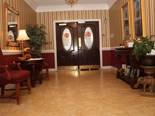 Interior view of an entrance area with double dark wooden doors featuring oval glass panels with stop signs. The room has beige tiled flooring, striped wallpaper with a maroon lower half, a small table with a lamp and flowers, two red cushioned chairs, a large mirror on the left wall, and a wooden console table with plants and decorative items on the right.