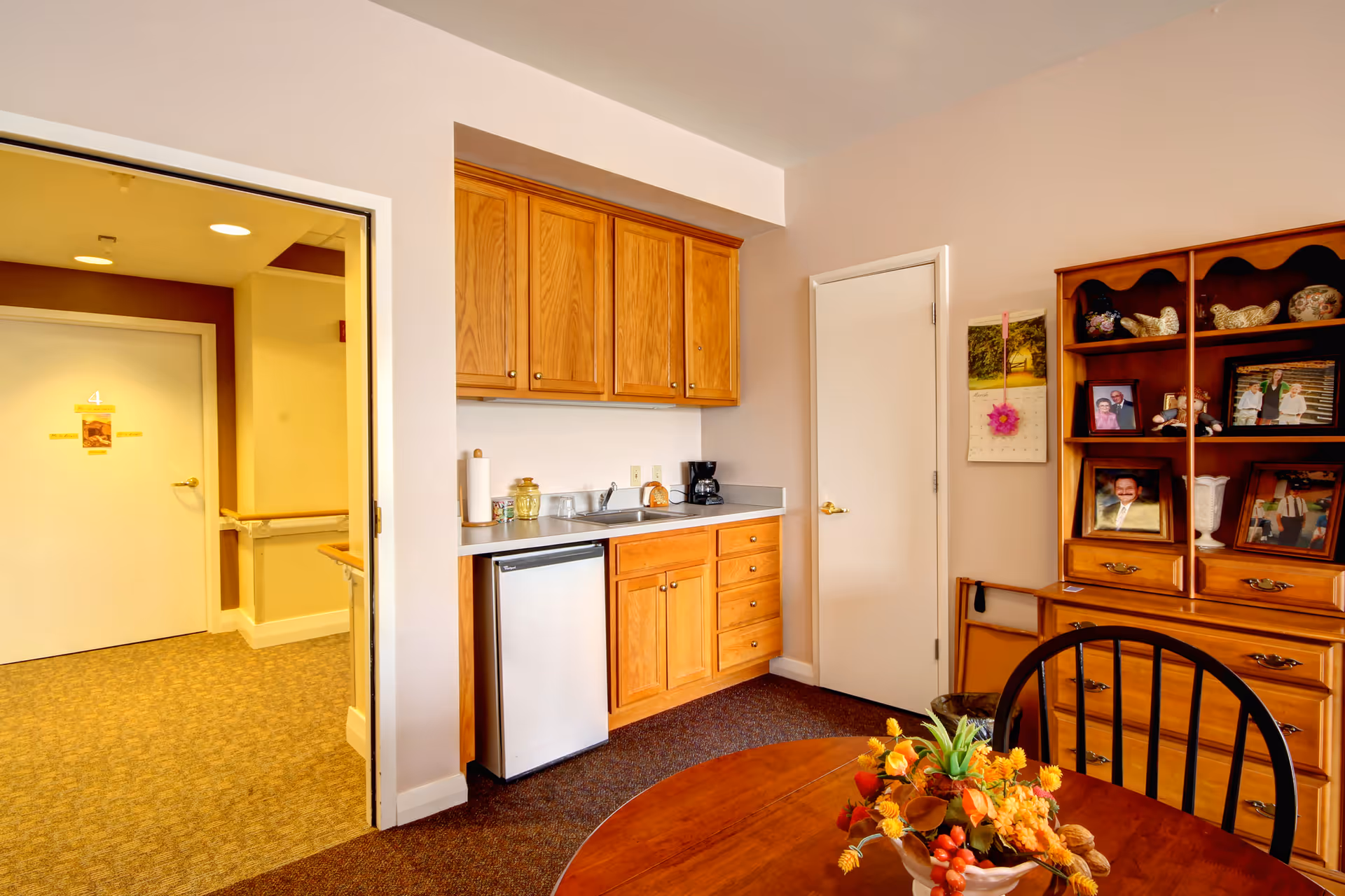 Interior view of a senior living facility room showing a small kitchenette with wooden cabinets, a mini refrigerator, a coffee maker, and a sink. To the right, there is a wooden hutch with framed photos and decorative items. In the foreground, a round wooden table with a floral centerpiece and a black chair are visible. An open door leads to a hallway with a white door labeled number 4.