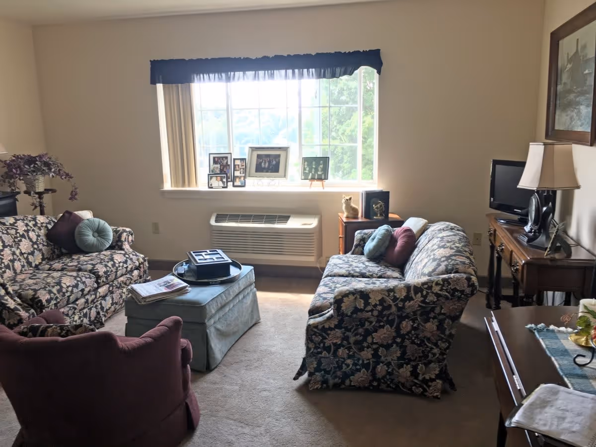 A cozy living room with floral patterned sofas, a maroon armchair, and a blue ottoman in the center. There is a large window with a blue valance letting in natural light. On the window sill are several framed photos. A small wooden side table holds a lamp and decorative items, and a TV sits on a wooden console table to the right. The room has beige walls and carpeted floor.