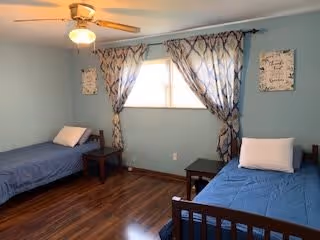 A bedroom with two single beds placed against opposite walls, each with blue bedding and a white pillow. There is a window in the center with patterned curtains, a ceiling fan with a light fixture above, and wooden flooring. Two small wooden side tables are positioned next to each bed. The walls are painted light blue and have decorative wall art on either side of the window.