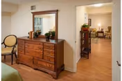 Interior view of a senior living facility room featuring a wooden dresser with a mirror, a chair with a round backrest, and a glimpse into a living area with armchairs and a lamp in the background.
