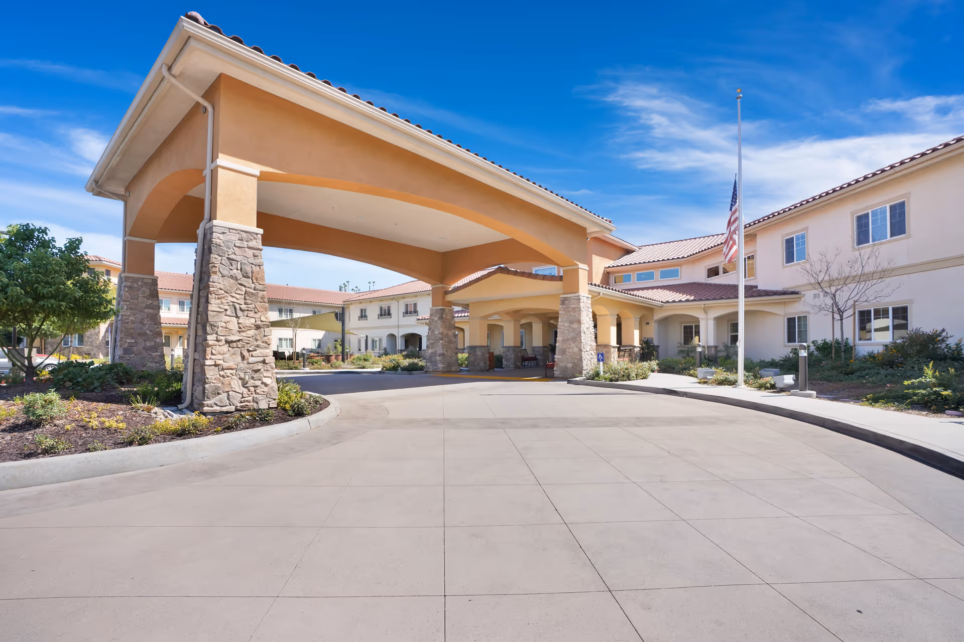 Front exterior view of a senior living facility with a large covered driveway supported by stone pillars, landscaped garden areas, and an American flag on a flagpole under a blue sky.