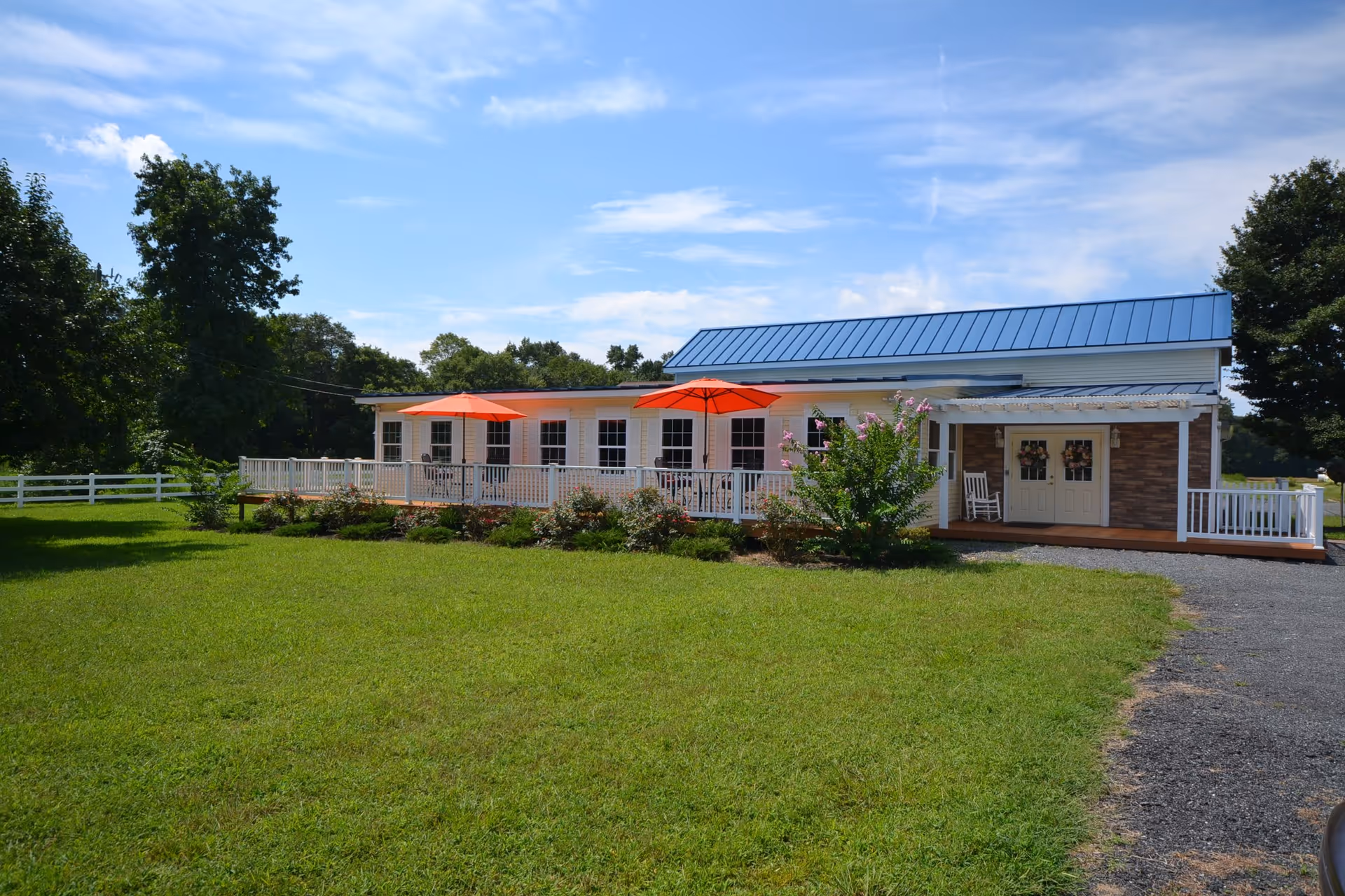 Exterior view of Chesapeake Cottage Assisted Living, a single-story building with a blue metal roof, white siding, and a porch with white railings. The porch has two orange umbrellas and outdoor seating. The building is surrounded by green grass, bushes, and trees under a partly cloudy blue sky.