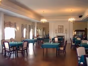 Spacious senior dining room with multiple tables covered in green tablecloths, wooden chairs, chandeliers, and a fireplace at the far end.