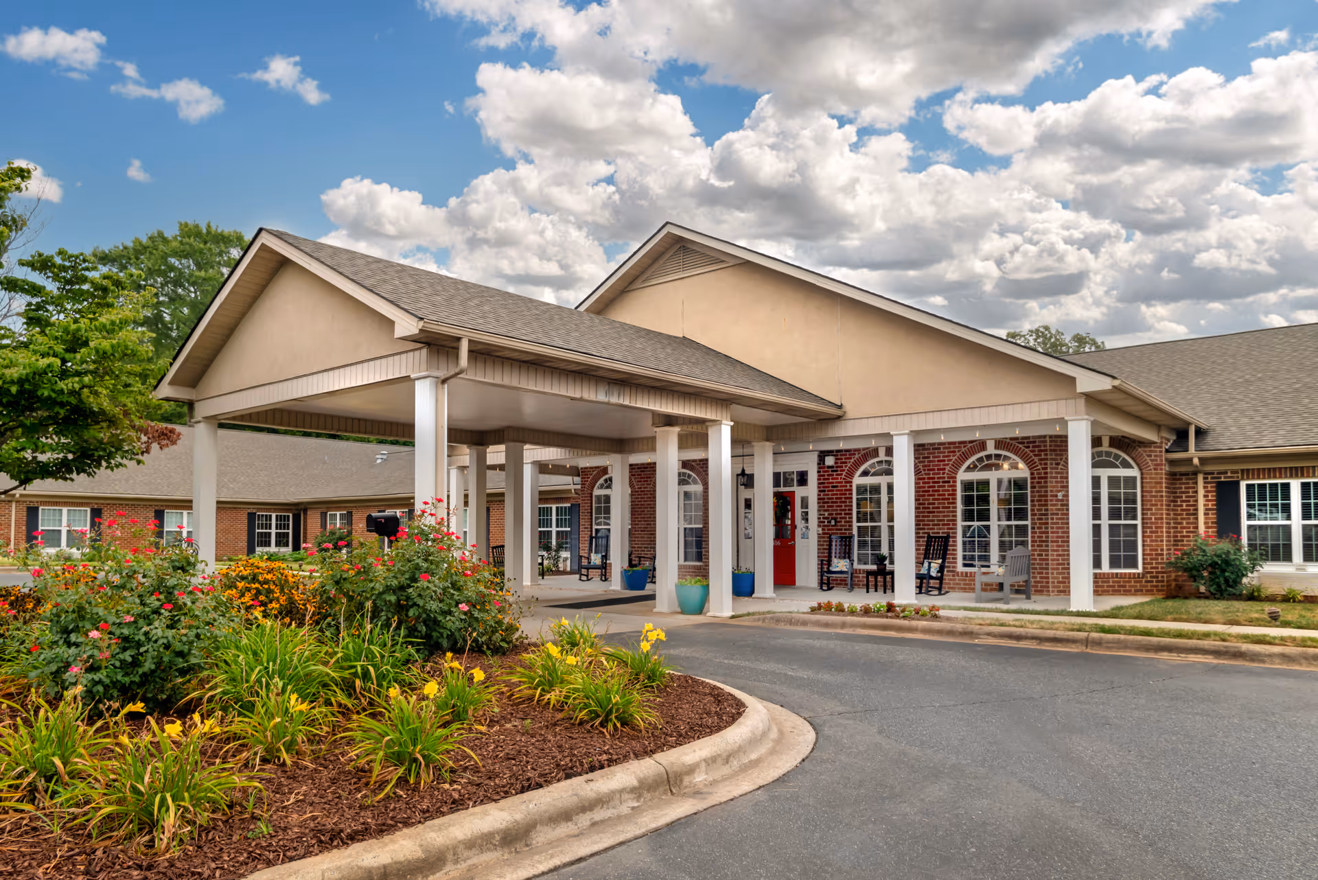 Exterior view of a senior living facility with a covered entrance supported by white columns. The building has a brick facade with large arched windows and a red door. There are colorful flower beds and greenery in front of the entrance, and the sky is partly cloudy.
