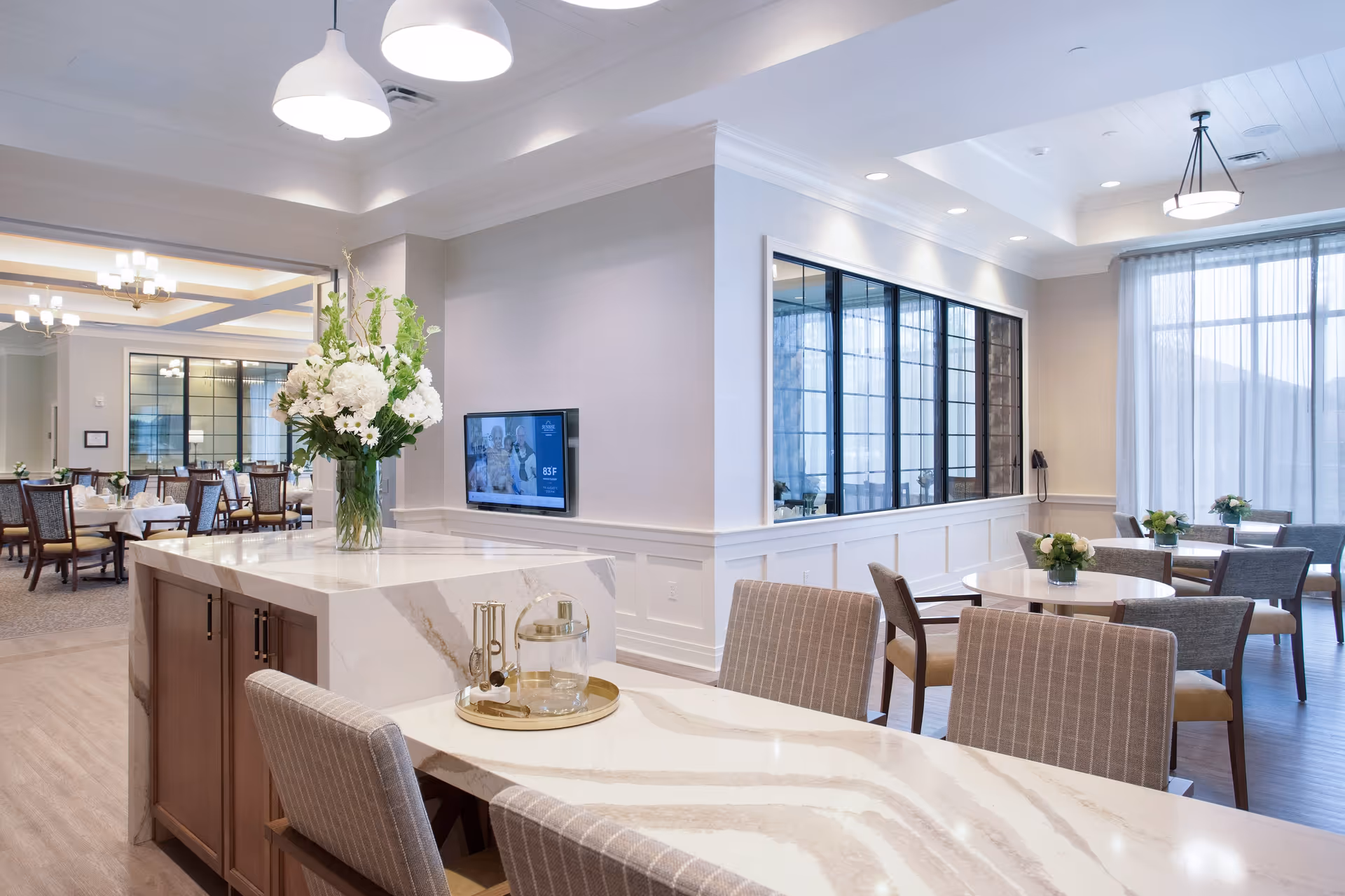 Bright, modern communal dining area with a marble-topped island, a vase of flowers, multiple dining tables and chairs, and large windows.