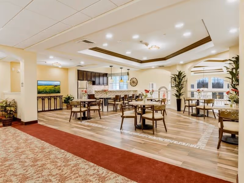 A bright and spacious dining area in a senior living facility with several round tables and chairs arranged on a wood-patterned floor. The room features a kitchen area with dark cabinets, a refrigerator, and a tiled backsplash. Large windows allow natural light to fill the space, and there are decorative plants and flowers on the tables. The ceiling has recessed lighting and a decorative tray design.